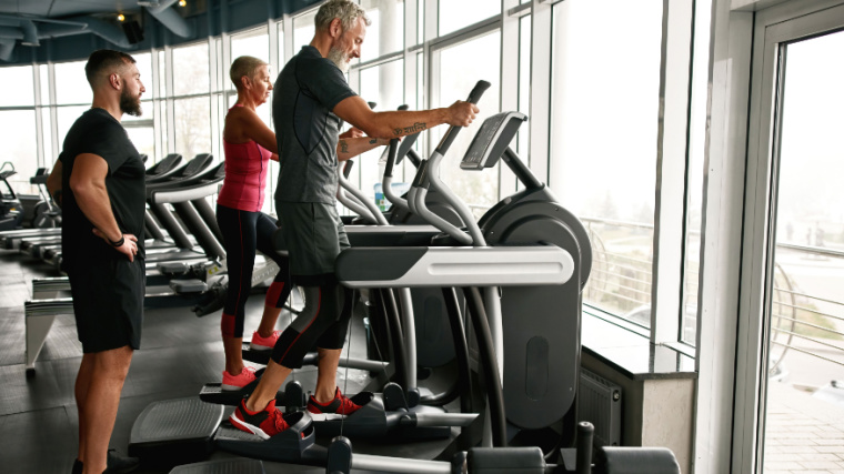 Two people doing using elliptical machines together with their personal trainer in the gym.