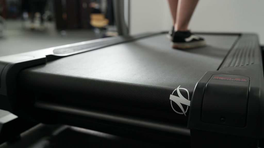A closeup of the the deck and belt of a NordicTrack X22i while someone is walking on it.