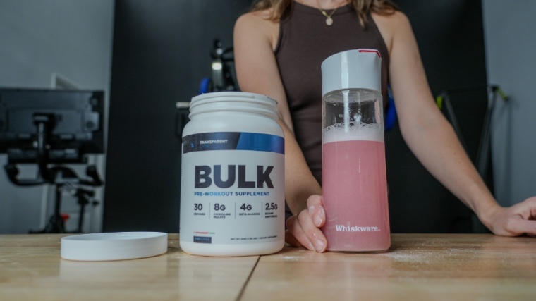 Container of Transparent Labs BULK Pre-Workout on wooden table beside woman holding shaker bottle.