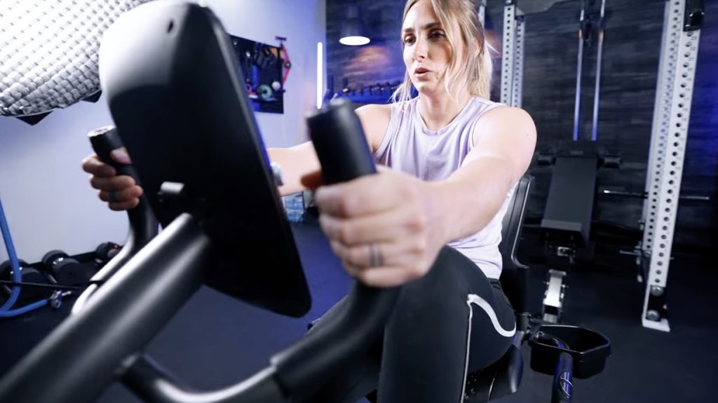 A woman is shown holding the handles while riding a Schwinn Recumbent exercise bike